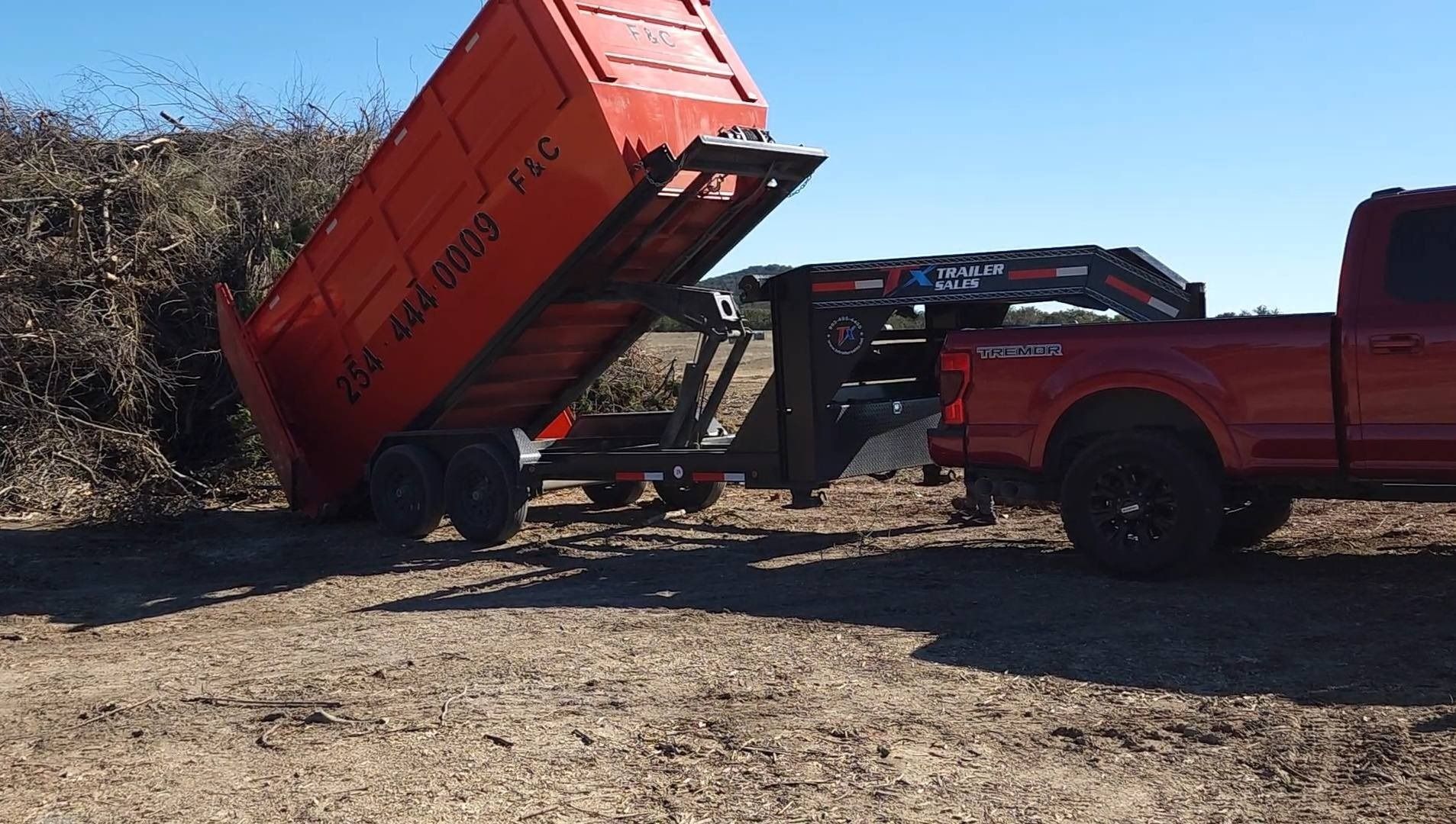 Red truck towing a dump trailer filled with brush, unloading onto the ground in a sunny outdoor setting.