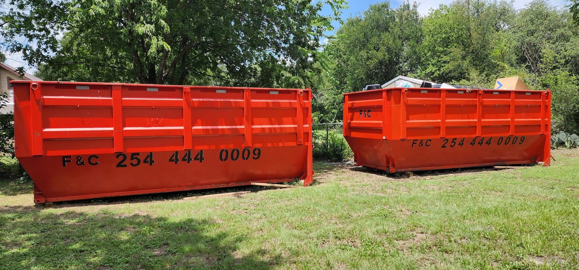 Two orange dumpsters on a grassy lawn with trees in the background, labeled with phone numbers.