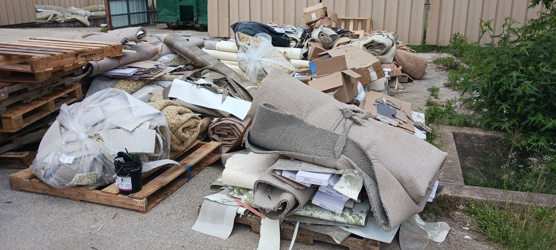 Pile of discarded materials including carpet, cardboard, and wood pallets. Located near green foliage and a wooden fence.