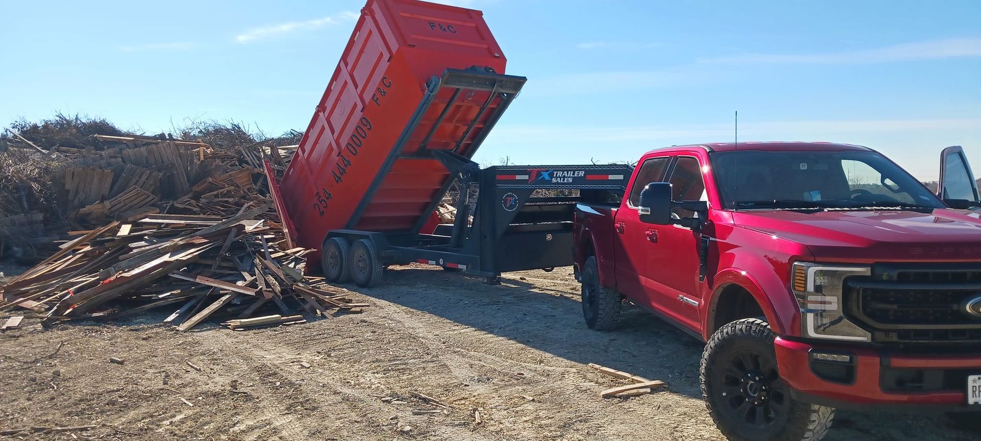 Red pickup truck with a dump trailer unloading wood debris on a sunny day.
