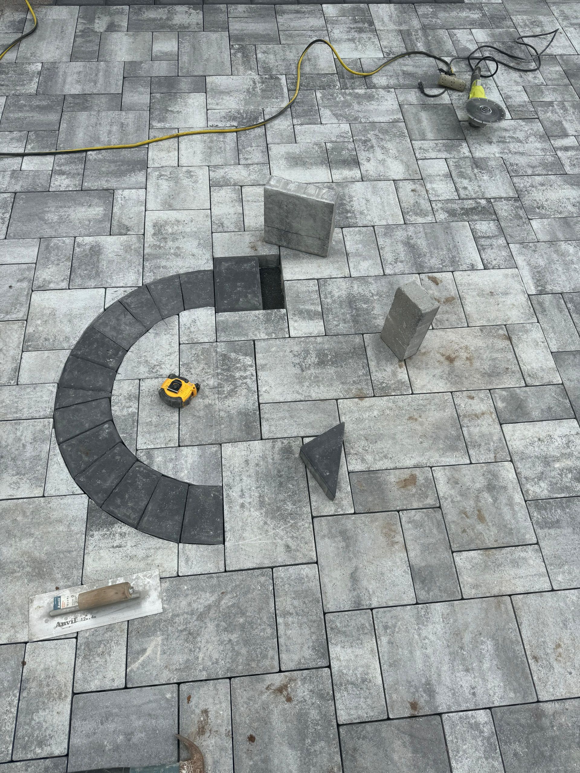 A semi-circle border of dark grey pavers set into a light grey stone patio, with tools and loose blocks nearby.