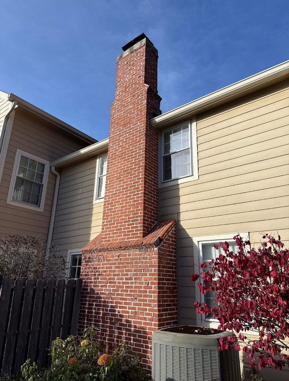 A tall brick chimney stands against a house with beige siding, a window, and a red-leaved shrub under a blue sky.