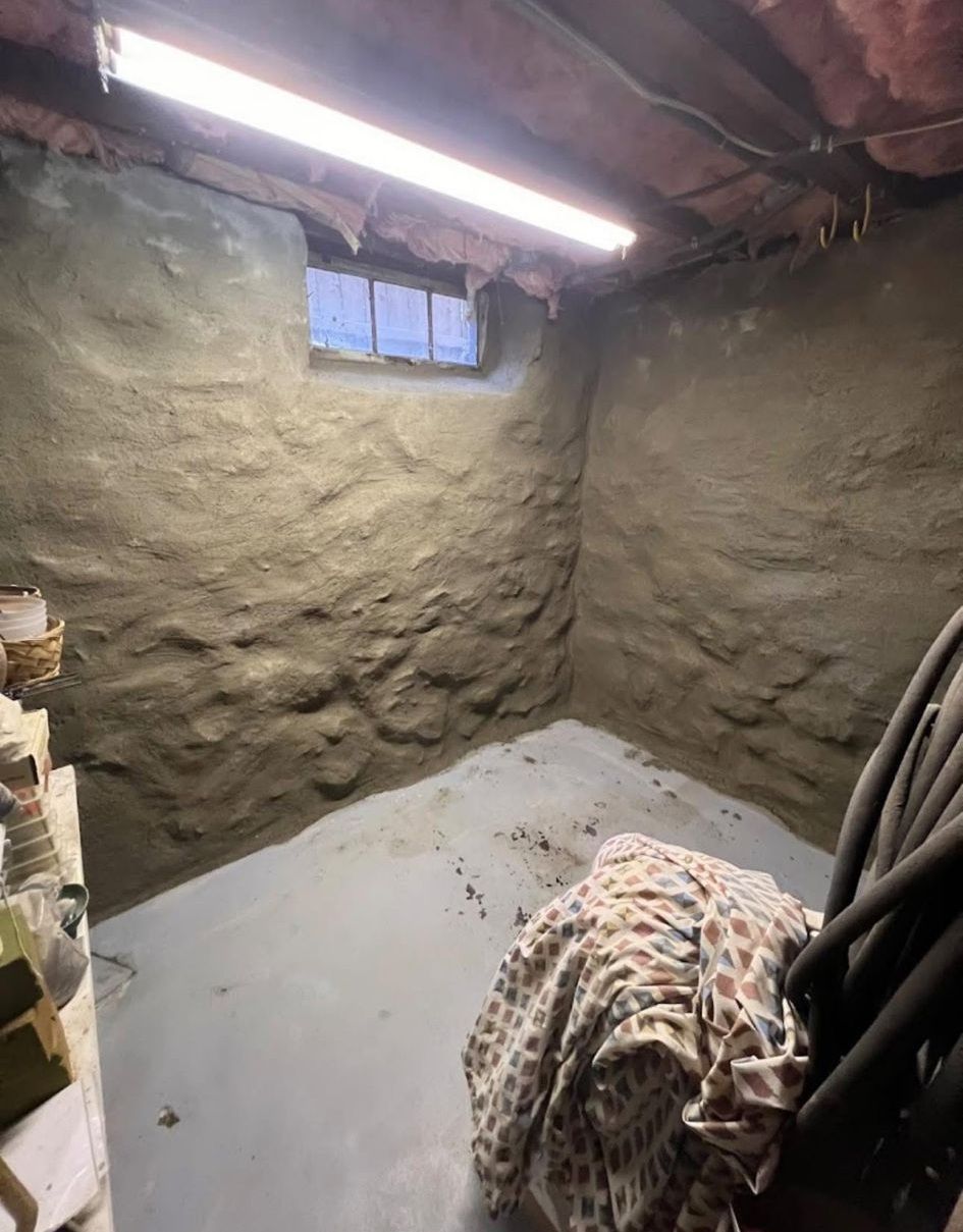 A basement corner with rough, parged stone walls, a small window, grey concrete floor, and overhead fluorescent light.