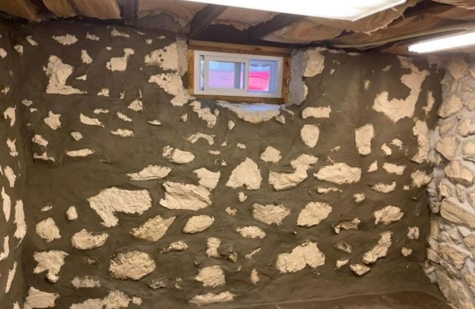 An unfinished basement wall featuring rough, light-colored stones embedded in dark gray mortar, with a small window above.