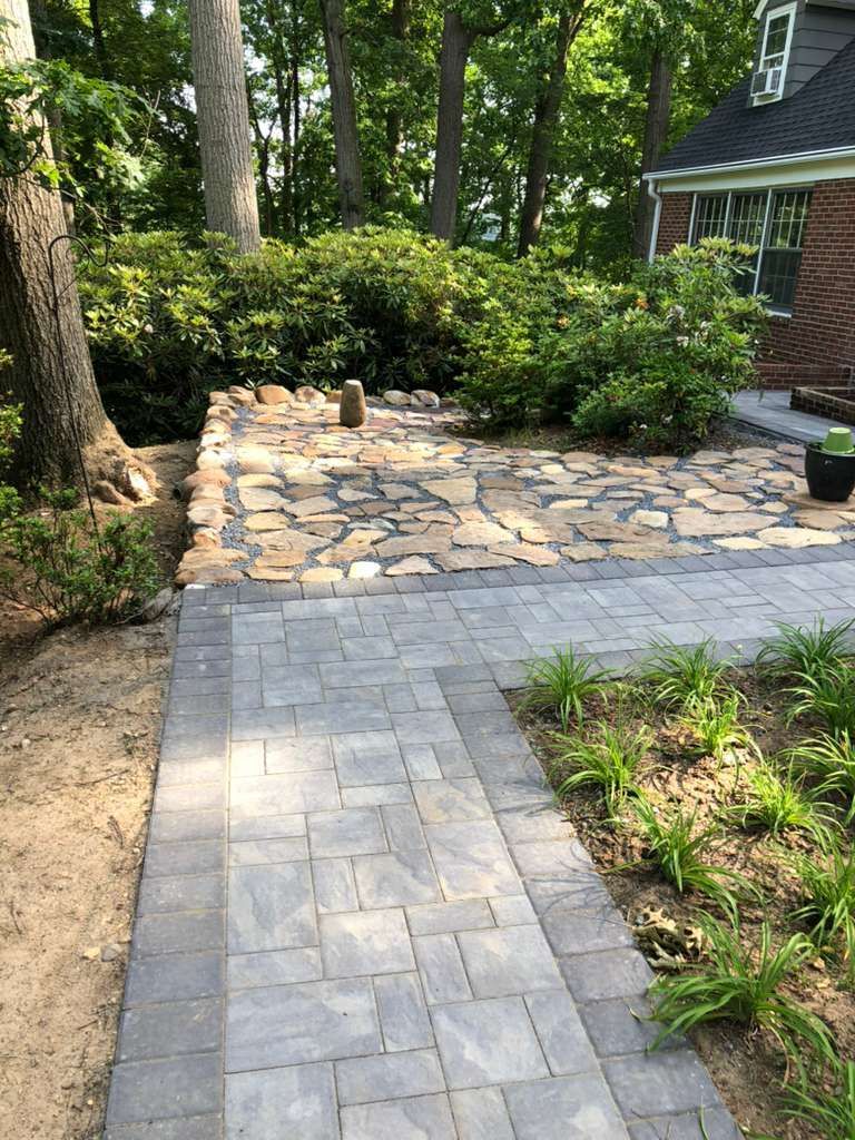 A paved stone walkway leads to a patio made of natural tan flagstones next to a brick house and lush green bushes.