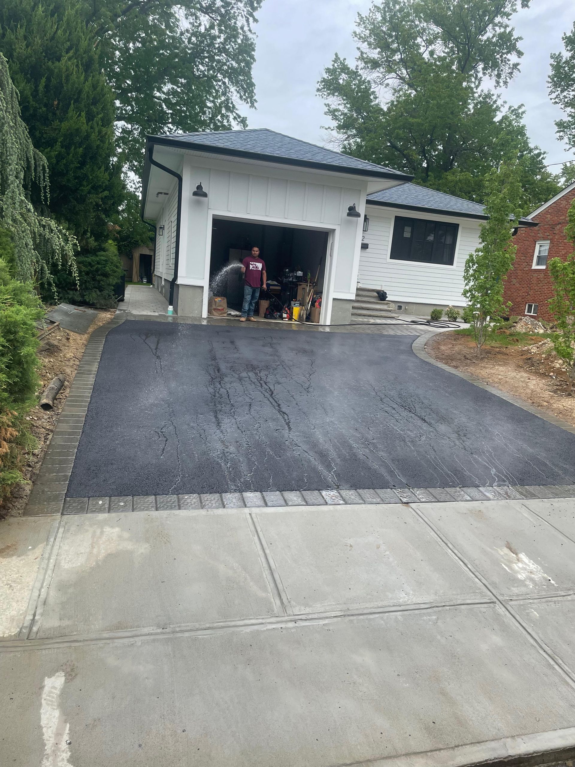 A freshly paved black asphalt driveway with a brick border leading to a white detached garage in a residential setting.