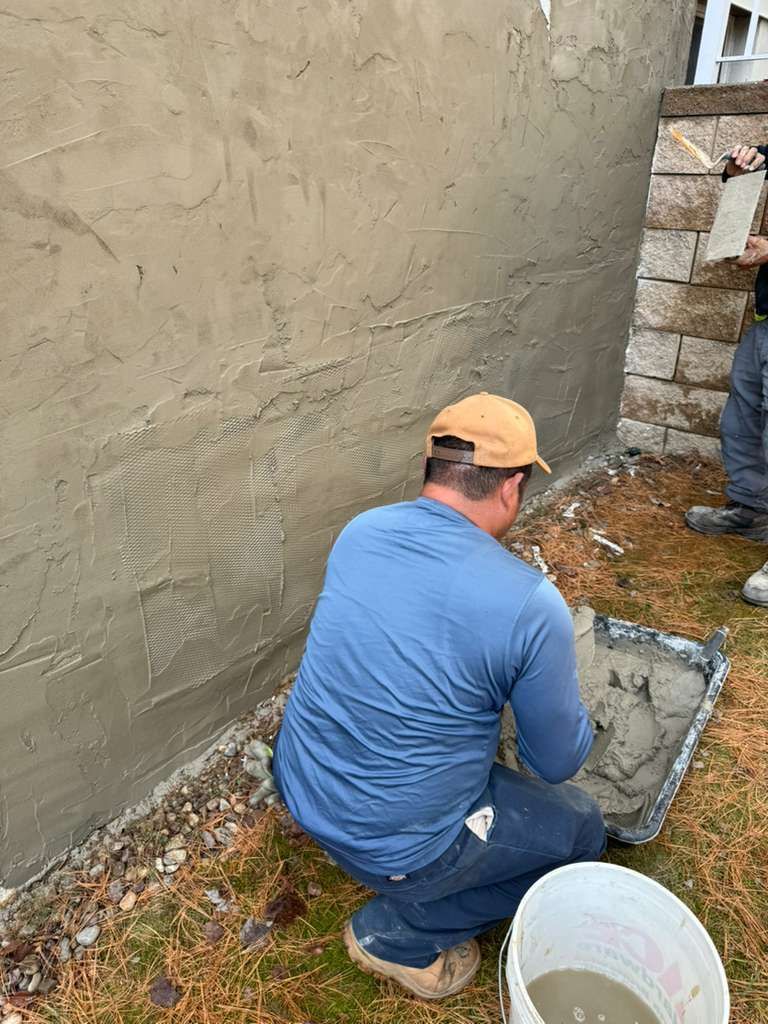 A person in a blue long-sleeved shirt and tan cap crouches while applying mortar to a textured wall near a bucket.