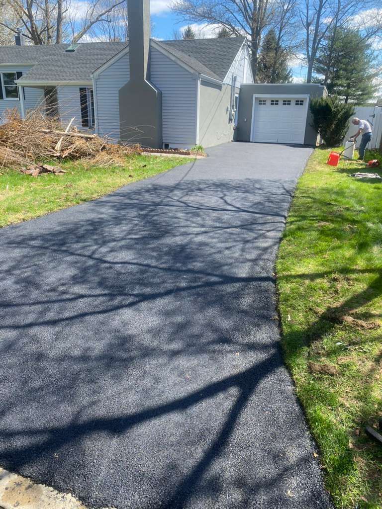 A newly paved black asphalt driveway leads to a gray suburban house with a white garage door on a sunny day.