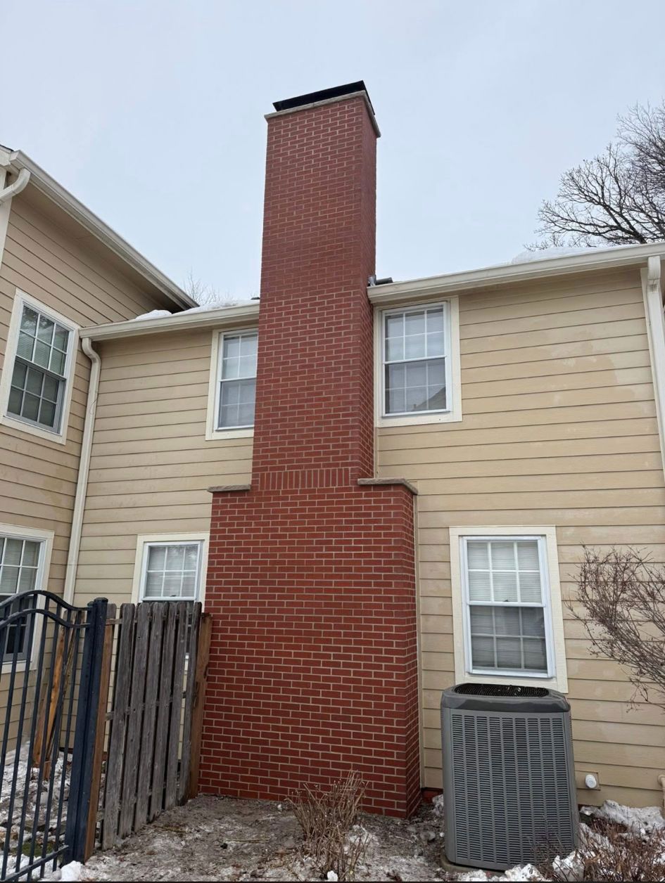A beige-sided house exterior featuring a prominent, rectangular red brick chimney stack rising from the ground.