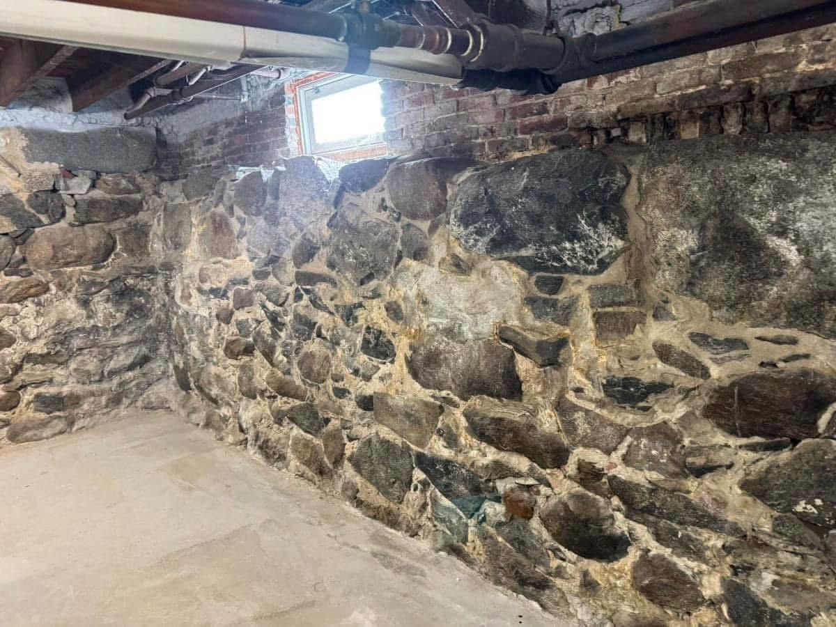 A corner of a basement featuring a rough stone foundation wall, a small window, and exposed pipes near the ceiling.