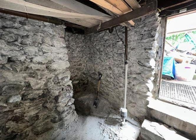 A stone foundation wall in a dimly lit basement, with a shovel leaning against the wall near a white pipe.