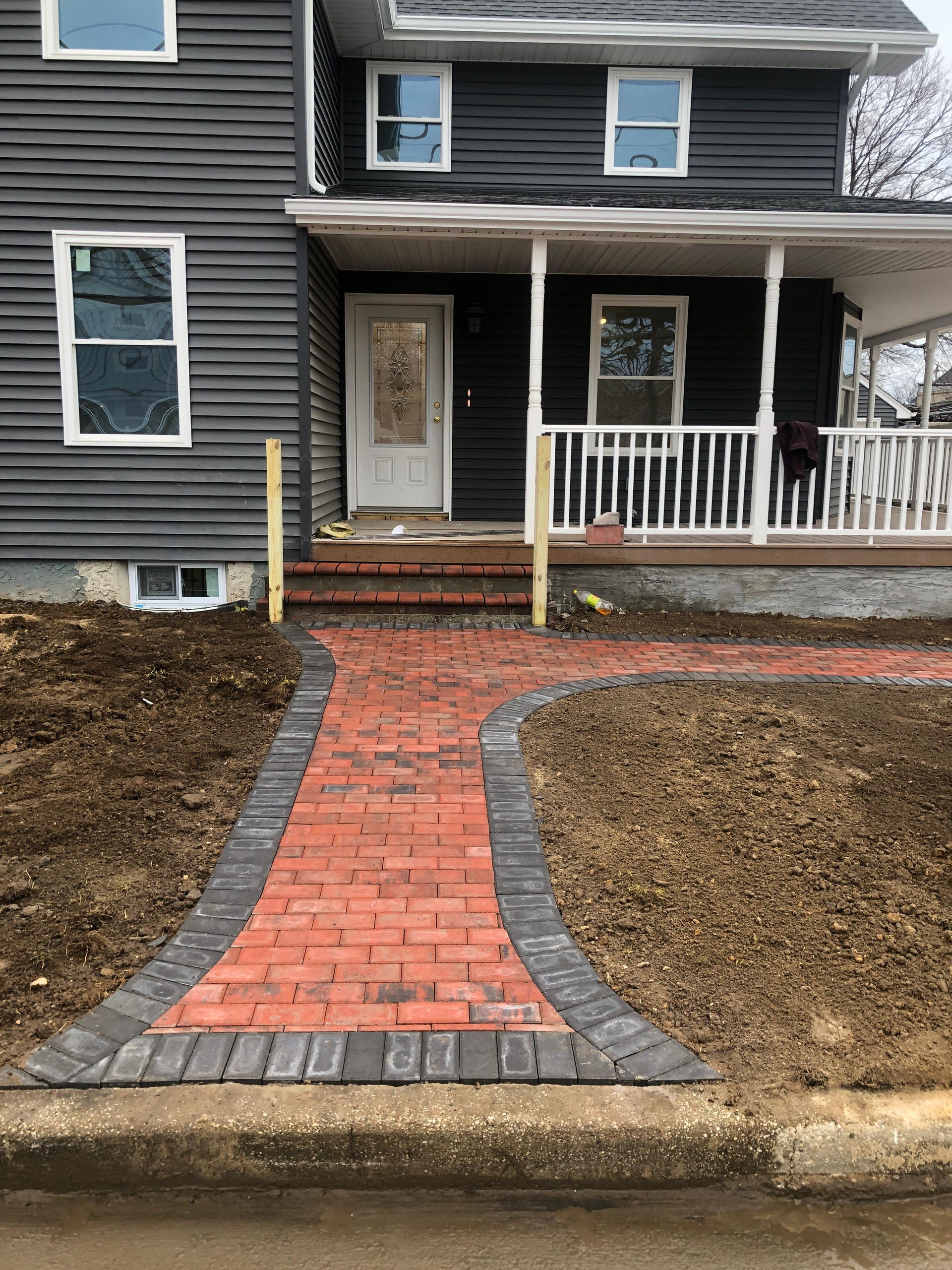 A newly laid red brick walkway with a dark border leads to the front porch steps of a dark-sided house.