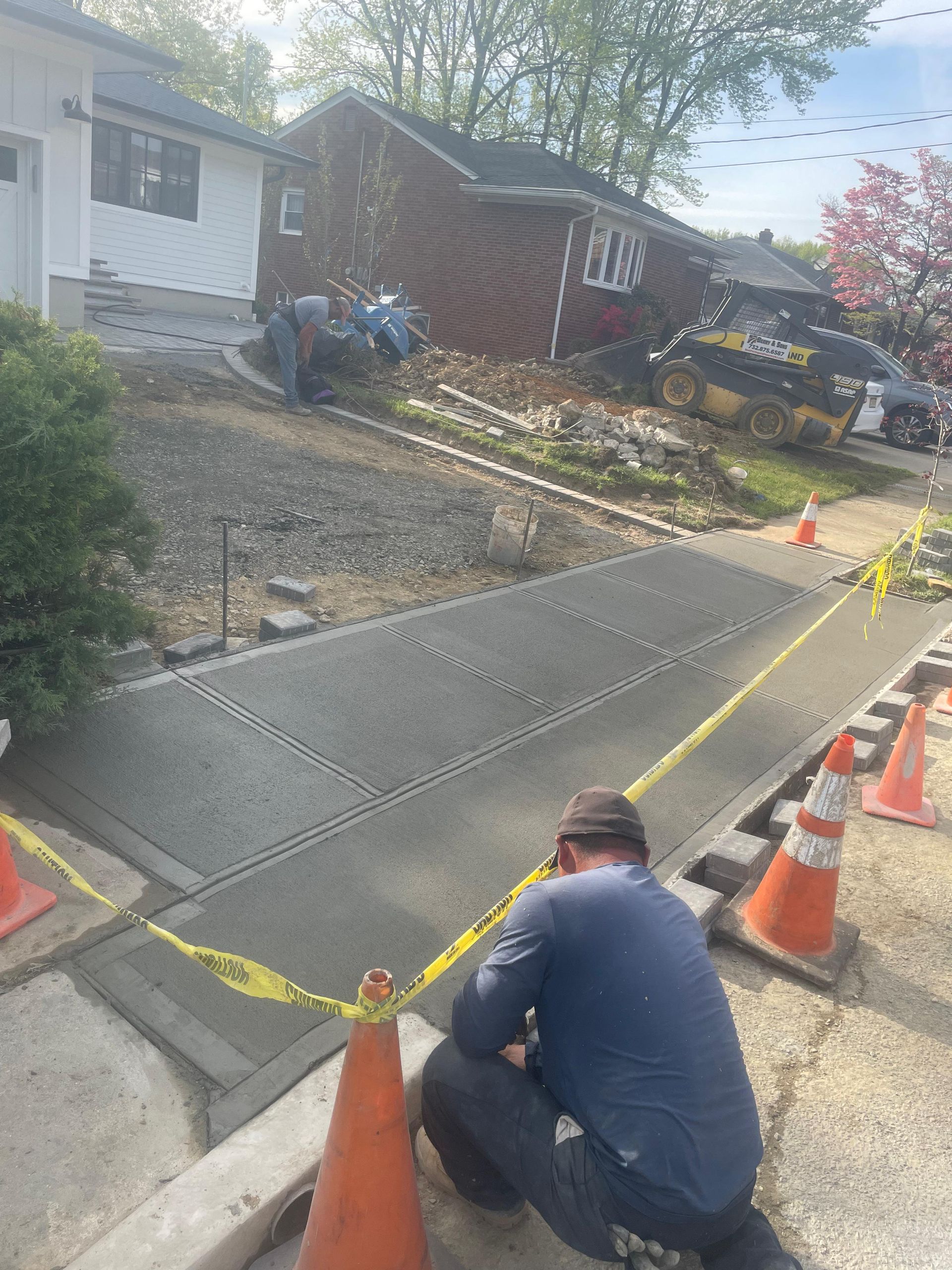 A person kneeling in a construction zone, finishing a newly poured concrete sidewalk bordered by orange safety cones.
