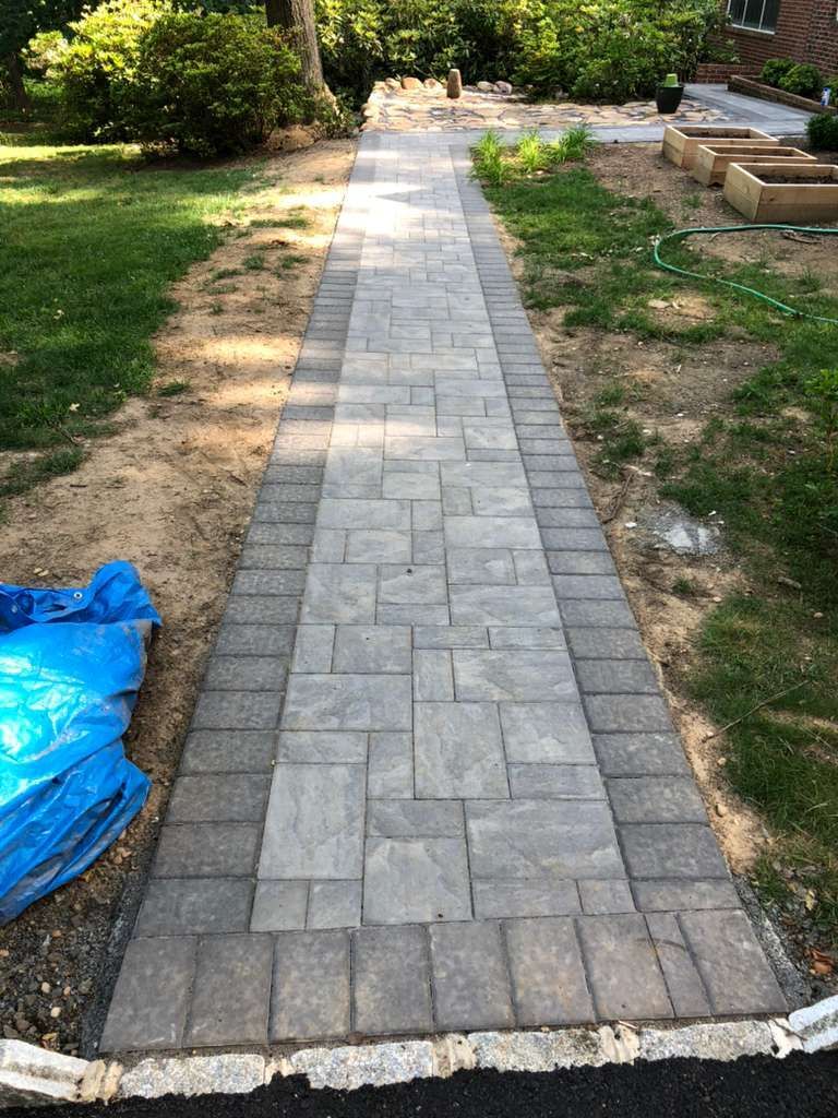A stone paver walkway with a dark border runs through a yard with green grass and garden beds.