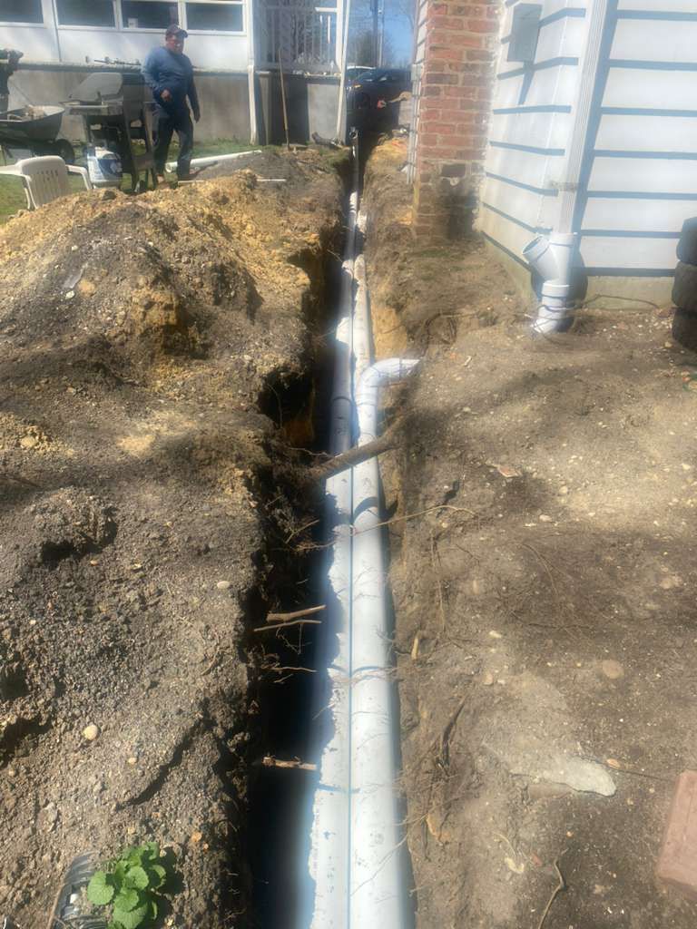 A construction worker in a deep trench installing a new grey PVC drainage pipe next to a brick building foundation.