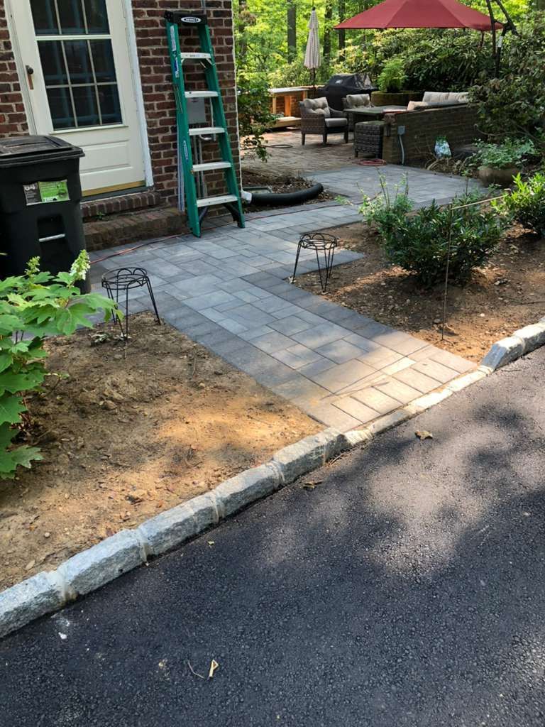 A paver walkway leads from an asphalt driveway to a brick house entrance, past garden beds and a green stepladder.