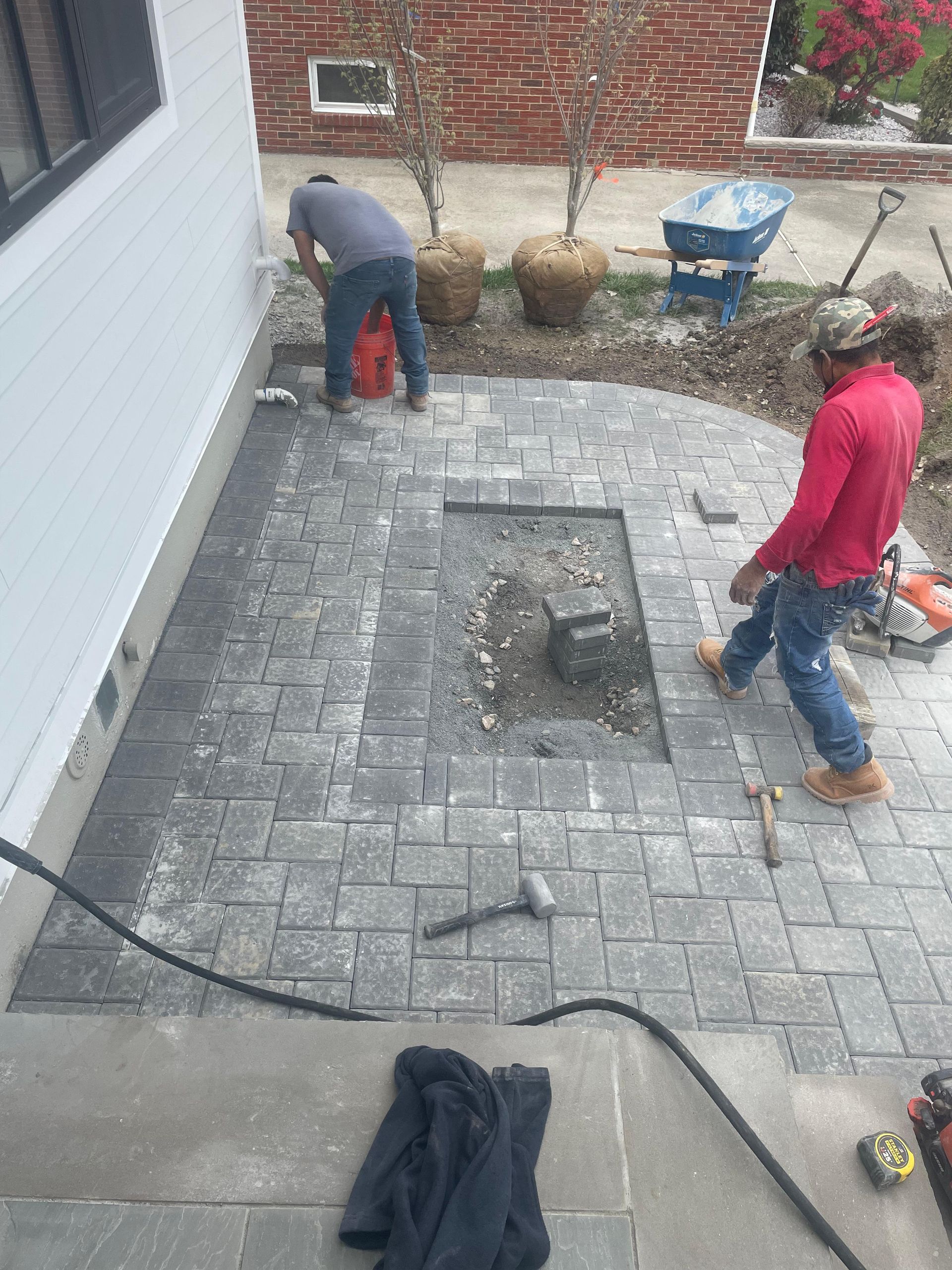 Two workers install a rectangular paver patio next to a house with a fire pit cutout in the center.