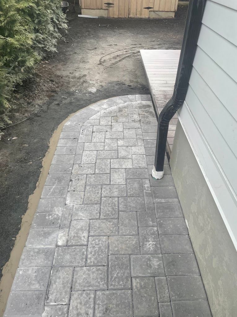 A grey paver stone walkway curves alongside the exterior wall of a house, adjacent to a wooden deck and soil.