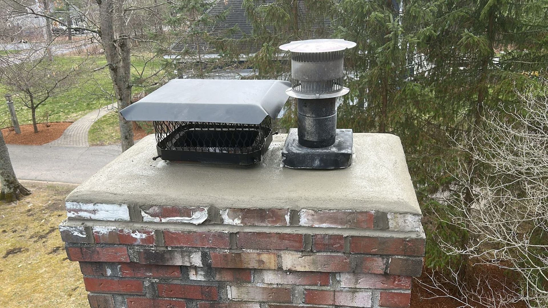 A brick chimney with a new concrete crown, topped by a rectangular metal flue cover and a circular chimney cap.