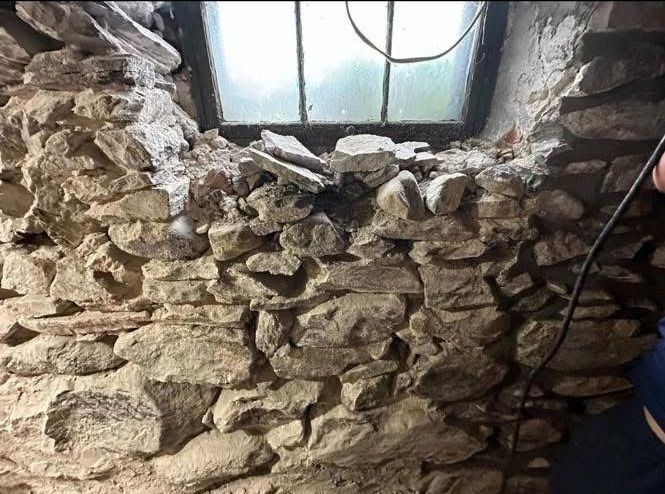 A crumbling stone basement wall under a window with loose rocks and mortar debris visible on the ledge.