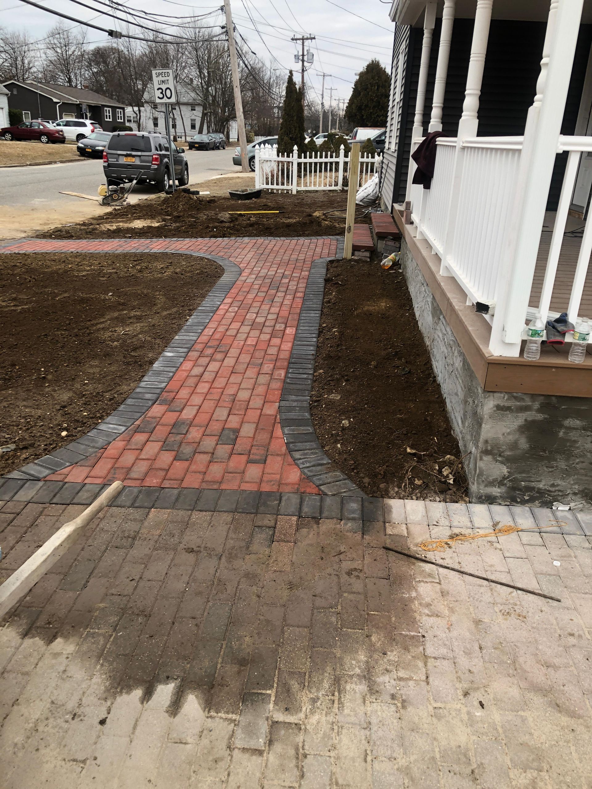 A red brick walkway with dark gray border stones leads toward a house porch, surrounded by patches of exposed brown soil.