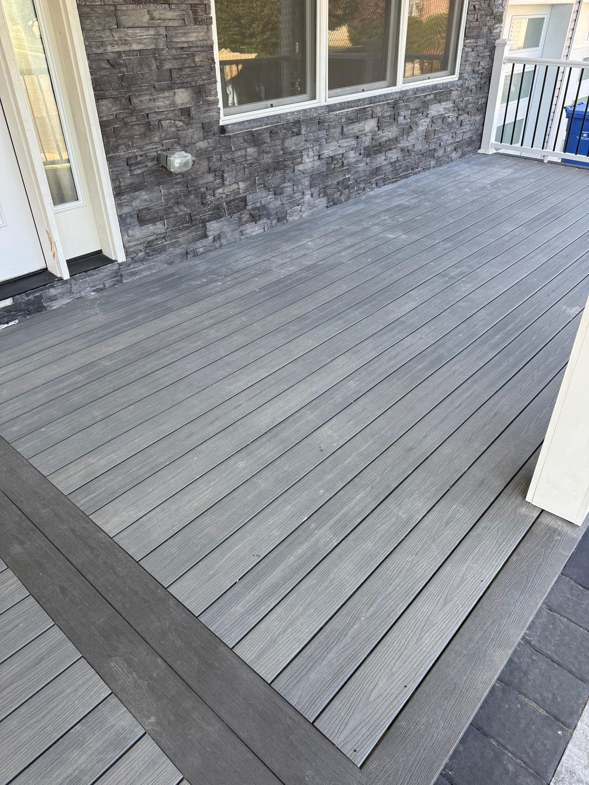 A gray composite deck with a darker gray border, adjoining a stone-faced house wall with windows and white trim.