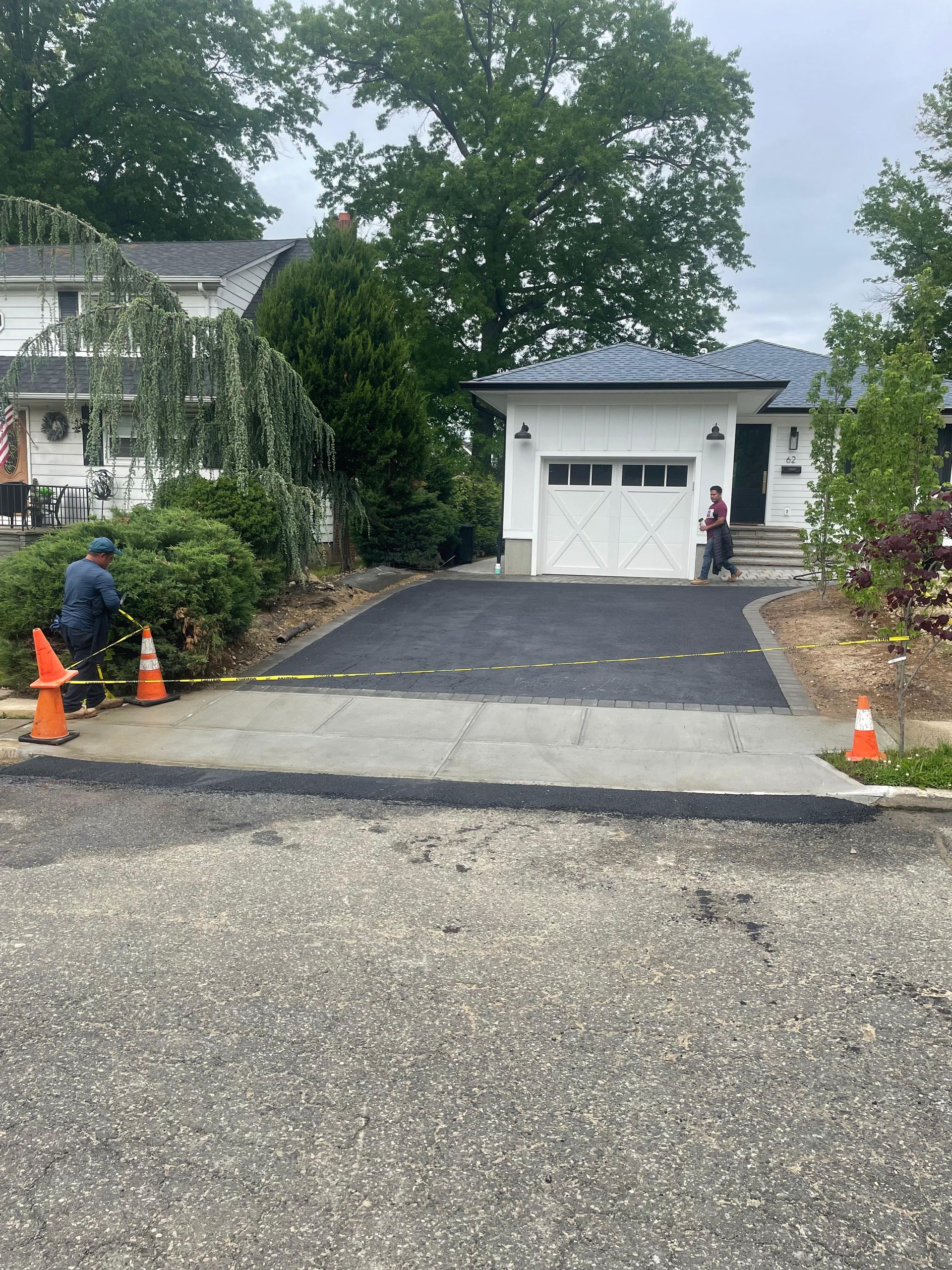 A person works on a newly paved driveway leading to a white detached garage near a house on an overcast day.