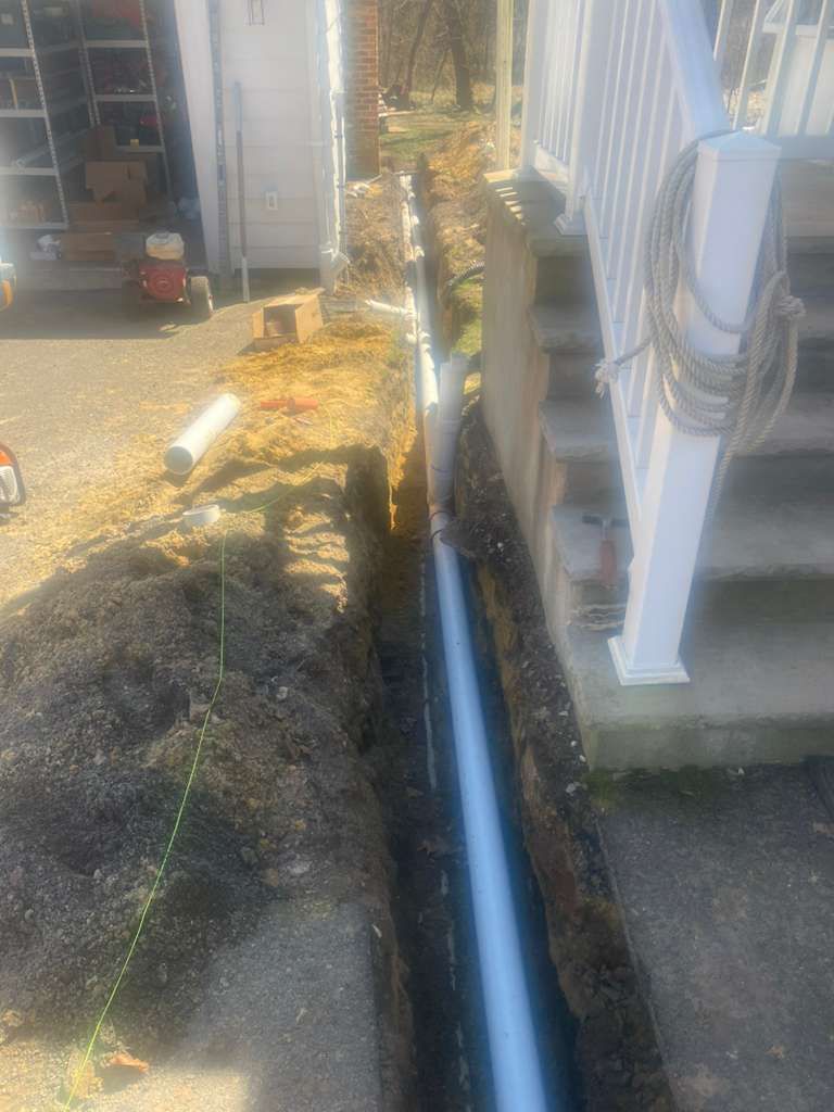 A trench dug along the side of a house with a new grey PVC pipe laid inside, next to a concrete staircase.