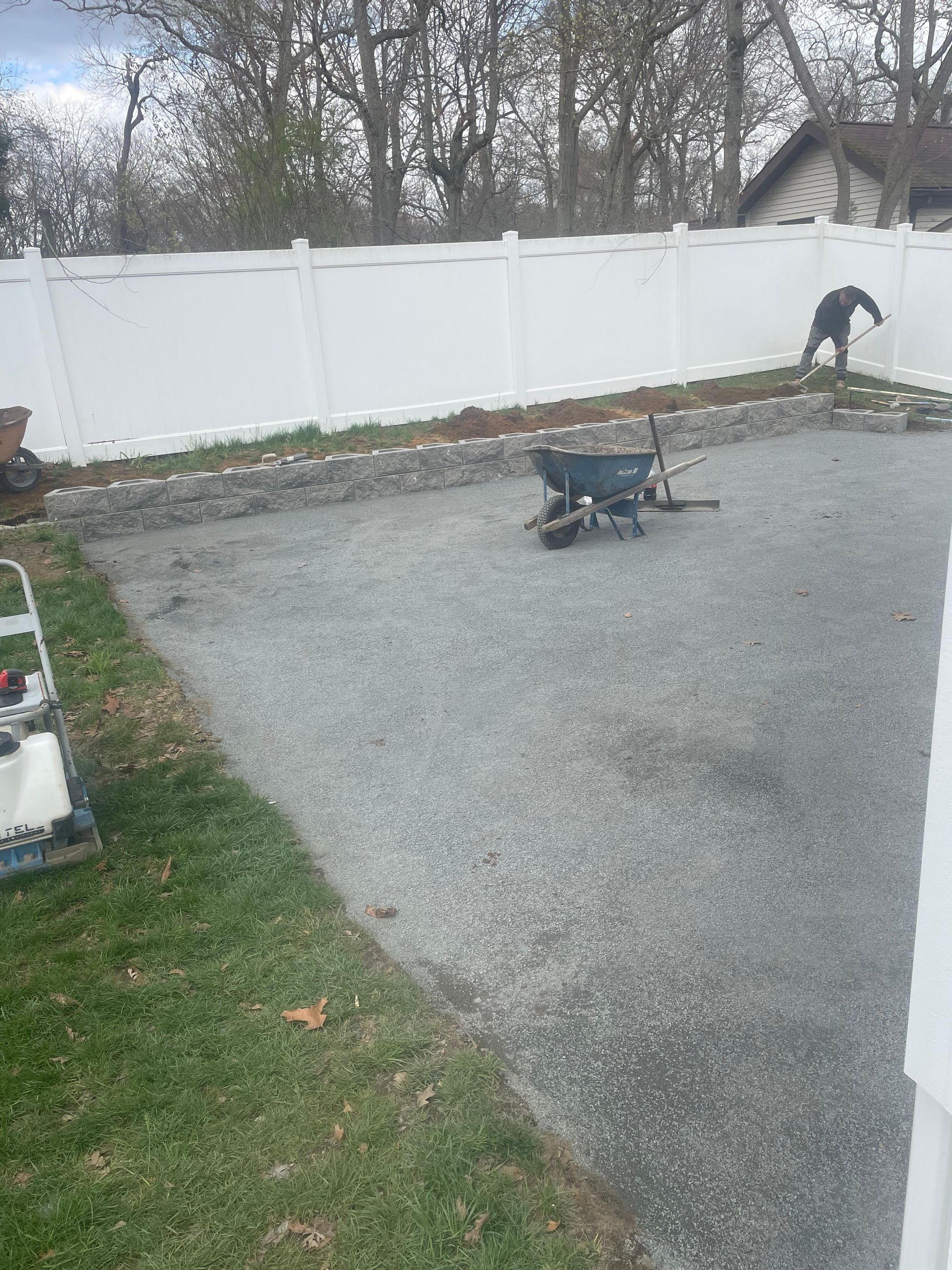 A person works in a residential backyard with a gravel area, a wheelbarrow, and a white privacy fence.
