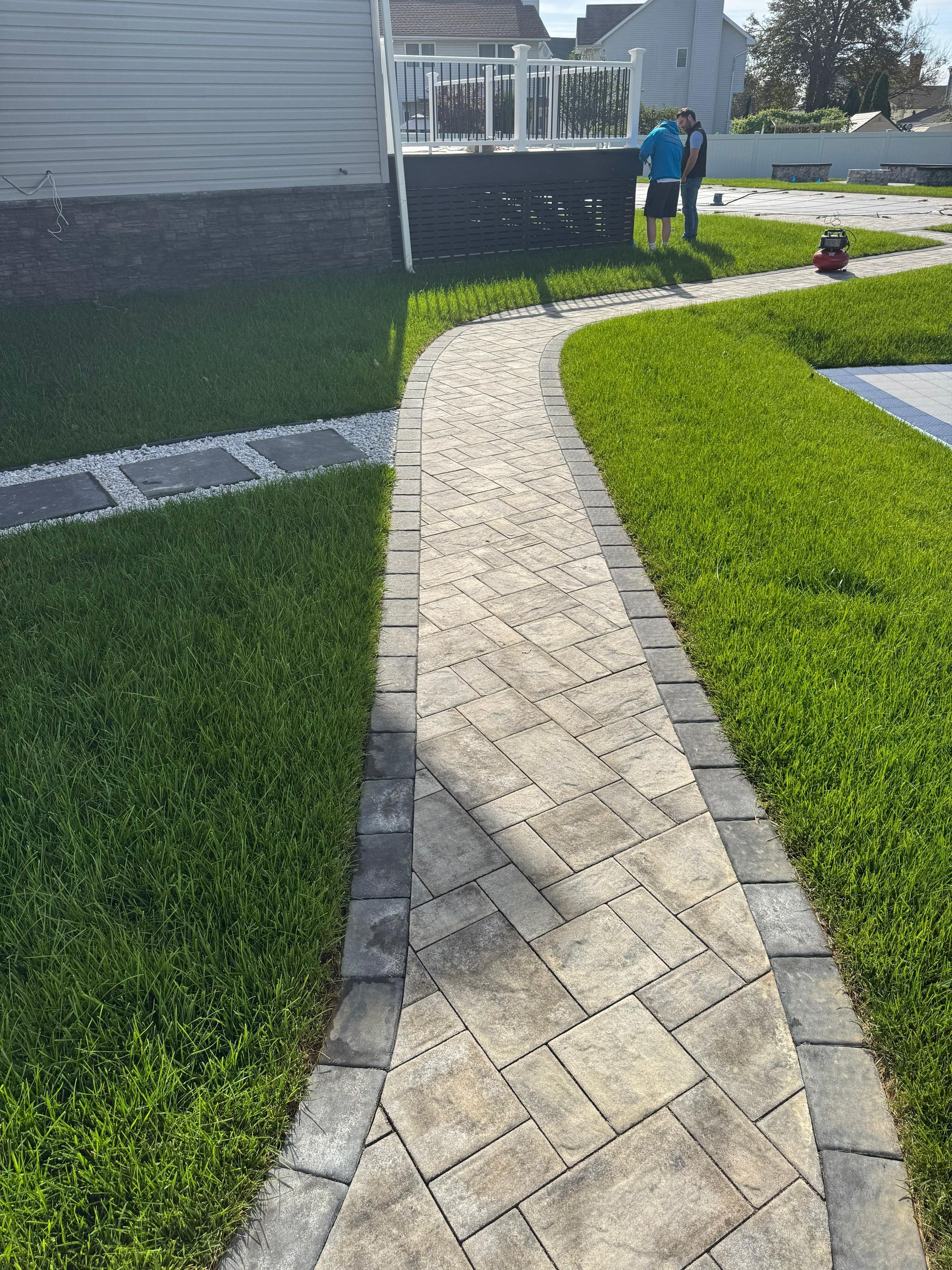 A light-colored stone paver path leads toward a house deck, flanked by dark stone borders and vibrant green grass.