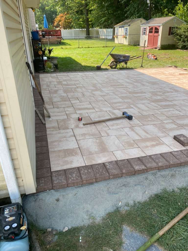 A freshly laid light-colored paver patio with a dark brick border, a mallet resting on top, set in a residential backyard.