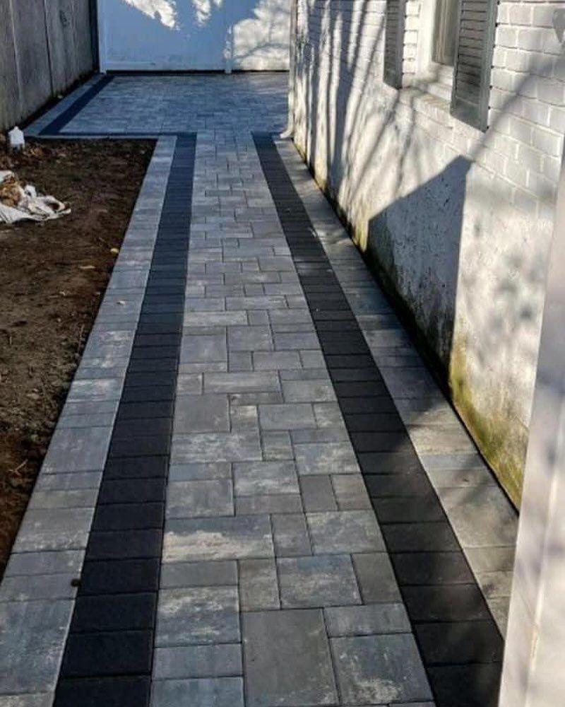 A newly installed stone paver walkway with a grey-toned center and dark border runs alongside a white brick house wall.