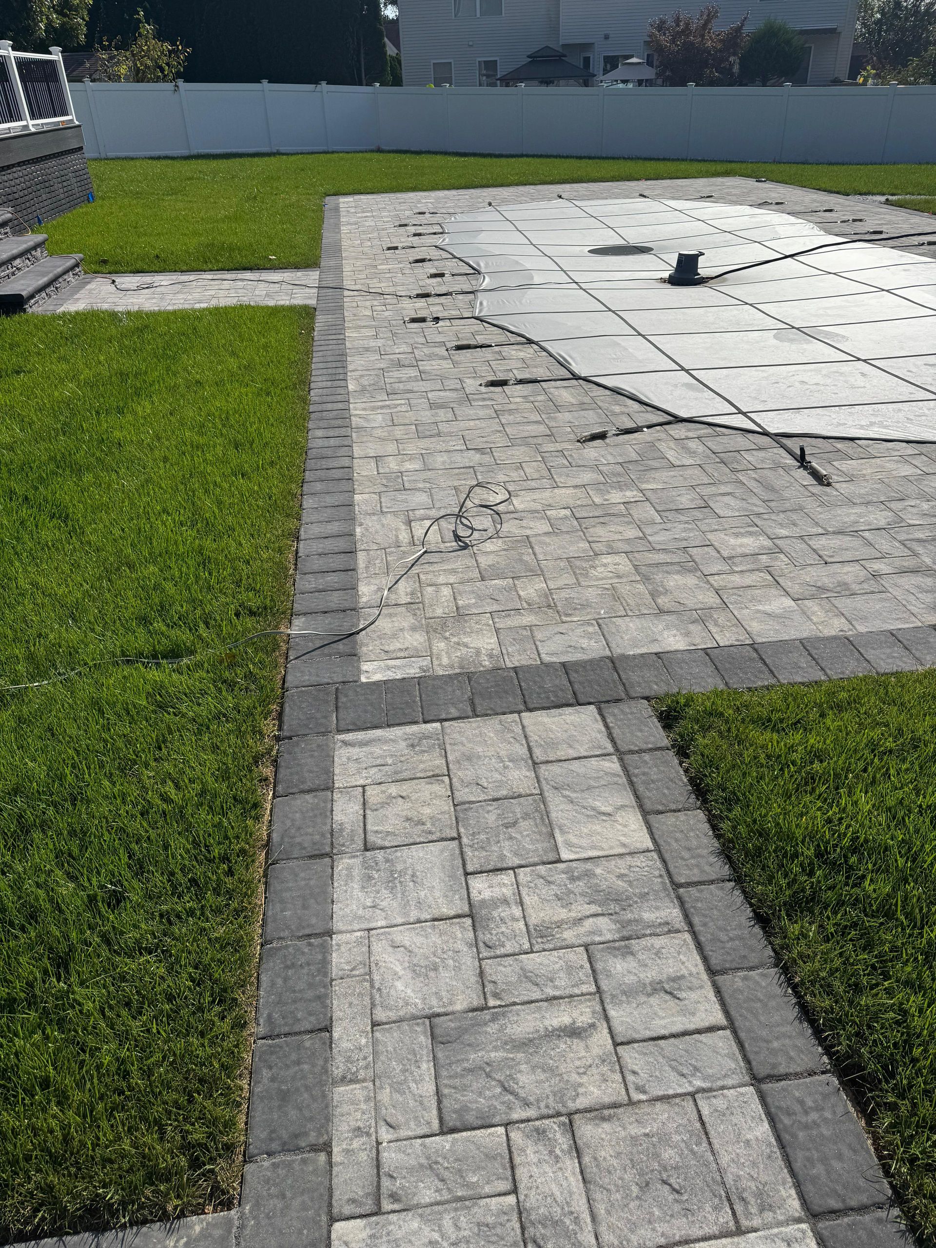 A paved stone walkway leads to a backyard swimming pool area with a white safety cover, surrounded by a green lawn.