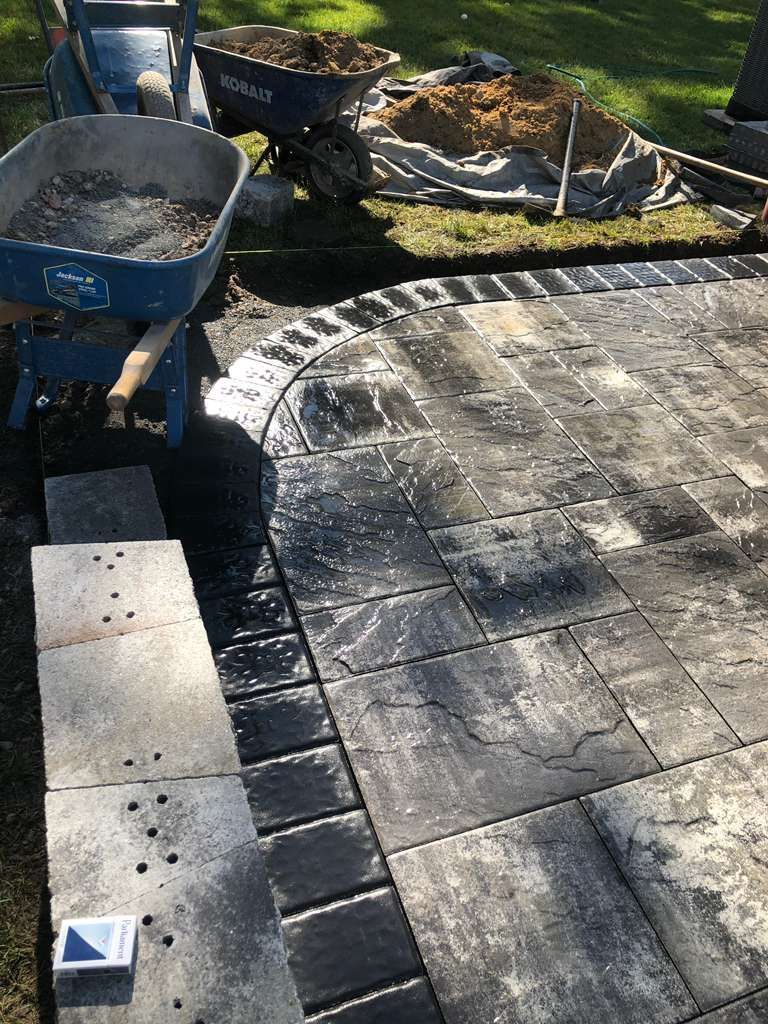A partially completed paver patio with a dark stone border and a stone wall in progress, with wheelbarrows in a yard.
