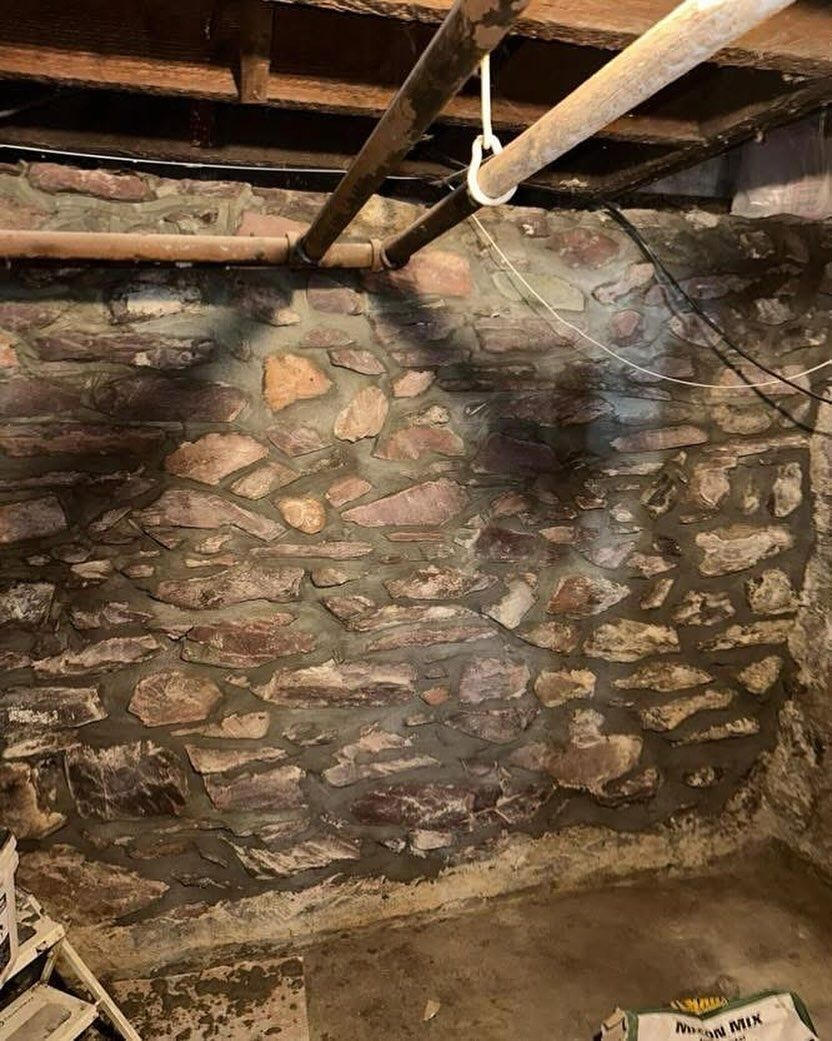 A basement wall constructed of irregular fieldstone and mortar, viewed from a low angle with overhead pipes.