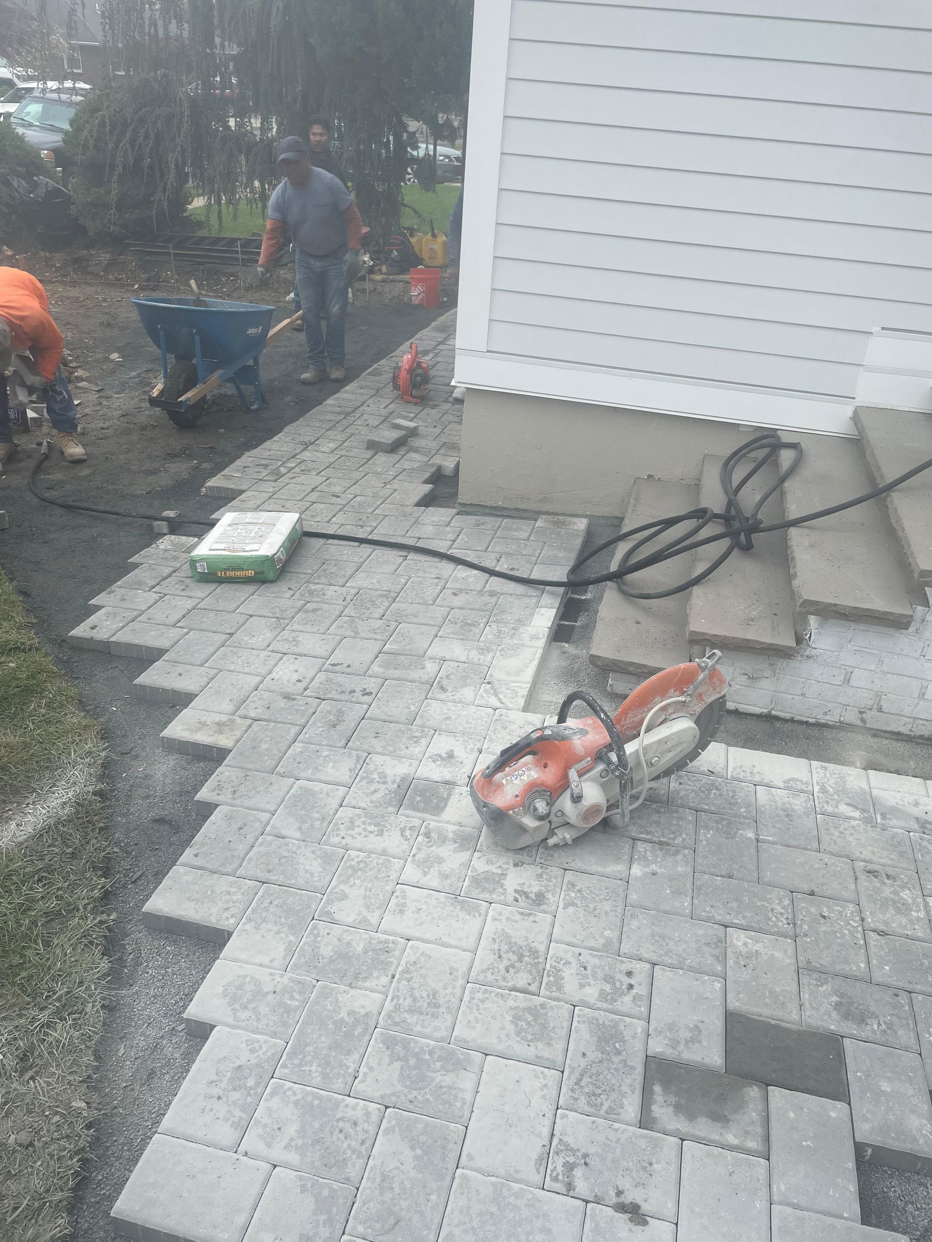 Two workers lay grey brick pavers on a walkway beside a house, with a masonry saw resting on the partially finished path.