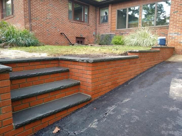 Red brick retaining wall and steps with dark stone caps leading to a grassy lawn in front of a house.
