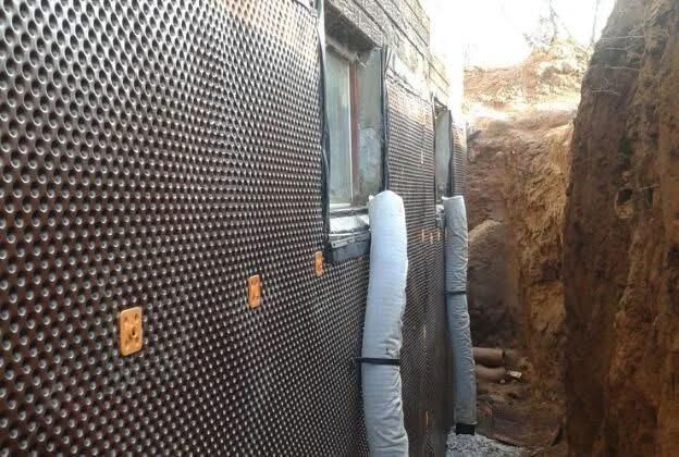 A foundation wall covered in dimpled drainage membrane and vertical drainage pipes, set within a deep, excavated trench.