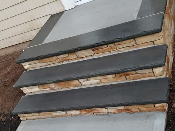 Three stone steps with dark slate treads and natural, tan-colored stone risers leading to a doorway.