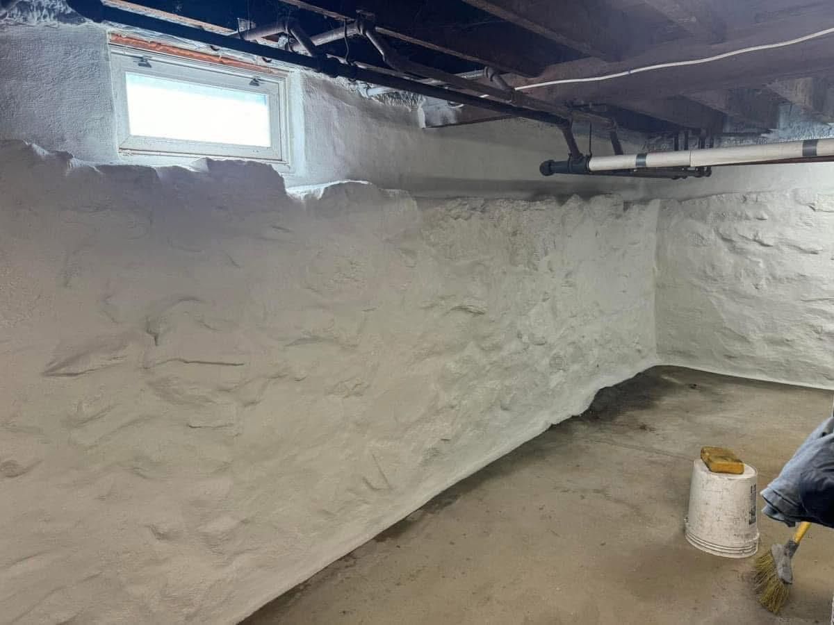 A white-painted, textured stone basement wall with a small window, exposed overhead pipes, and a bucket on the floor.