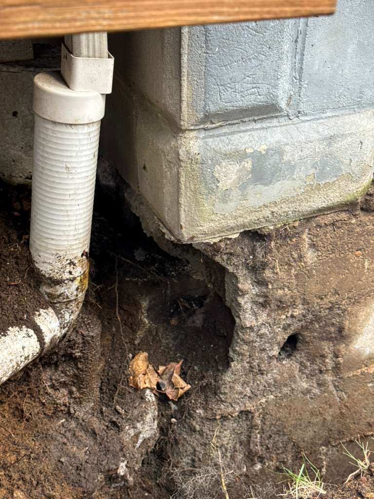 A white downspout positioned next to a section of a foundation wall with visible soil erosion and a gap beneath the base.