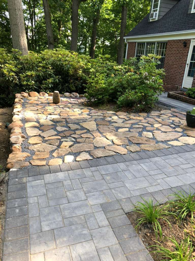 A stone patio made of irregular natural rocks bordered by rectangular grey pavers, set in a yard next to a brick house.