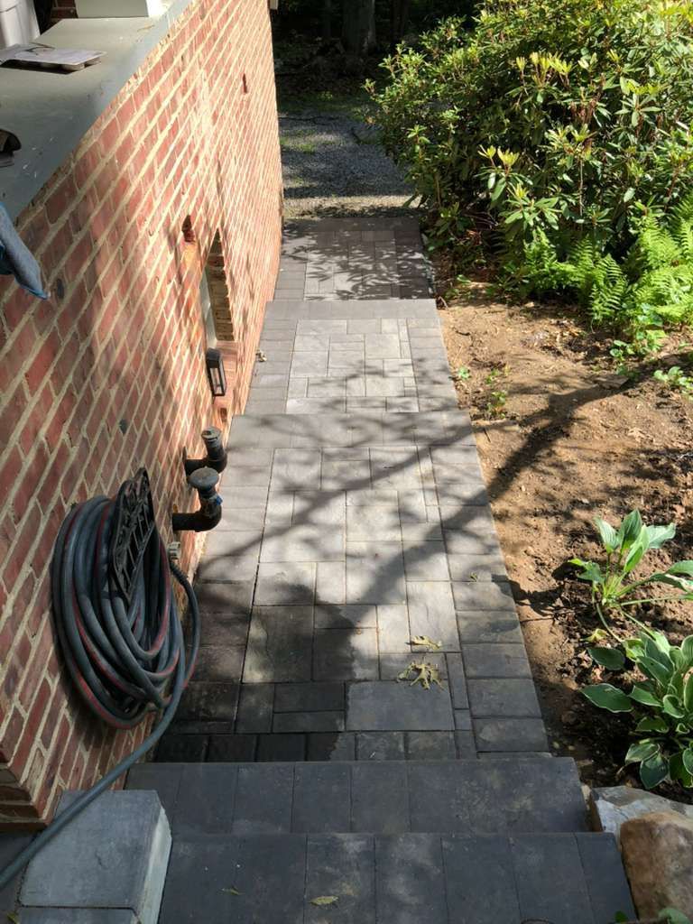 A downward-sloping outdoor staircase made of dark gray pavers alongside a brick wall and a garden area.