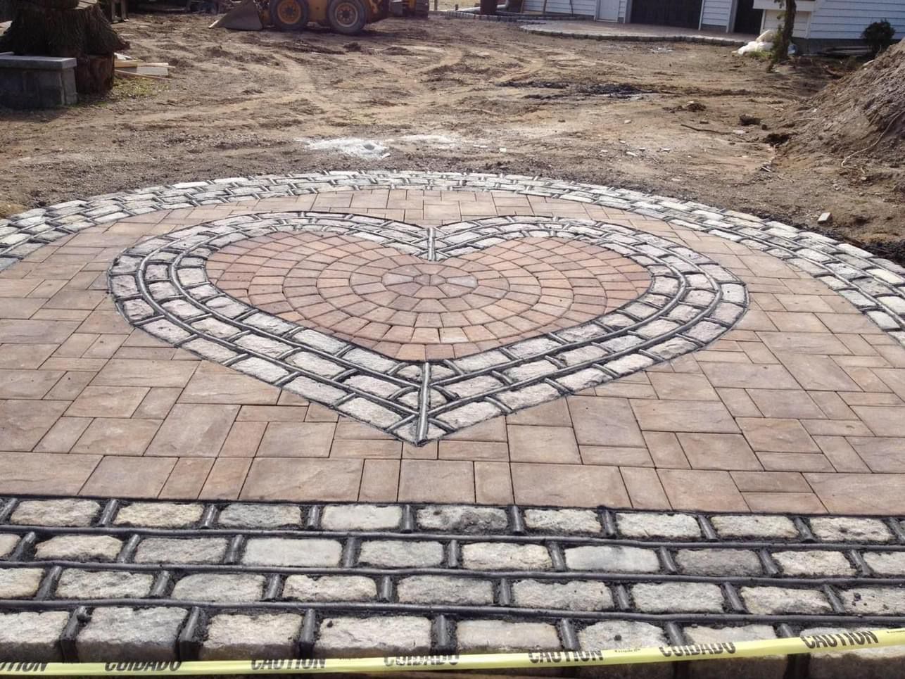 A circular stone patio under construction featuring a heart-shaped brick inlay surrounded by gray cobblestone borders.
