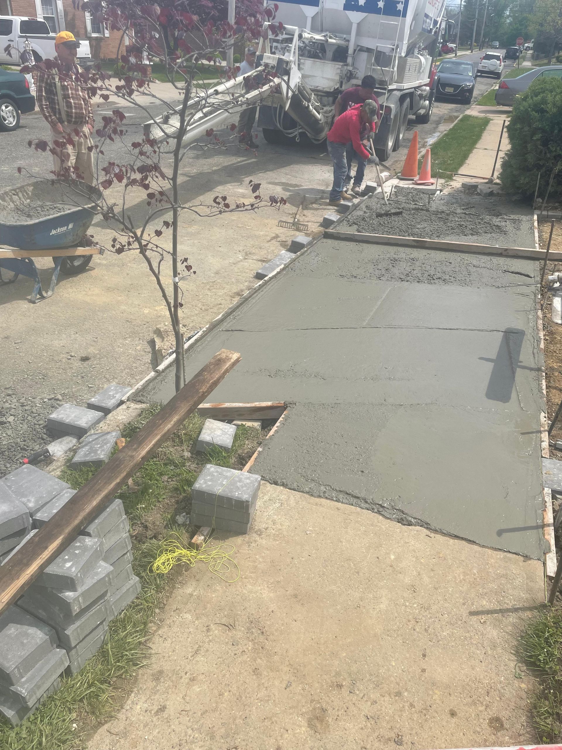 Construction workers pour fresh concrete to form a new sidewalk section next to a truck and stacks of paving stones.
