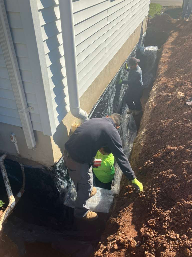 Two workers install waterproofing material along the exterior foundation of a house in a deep trench.