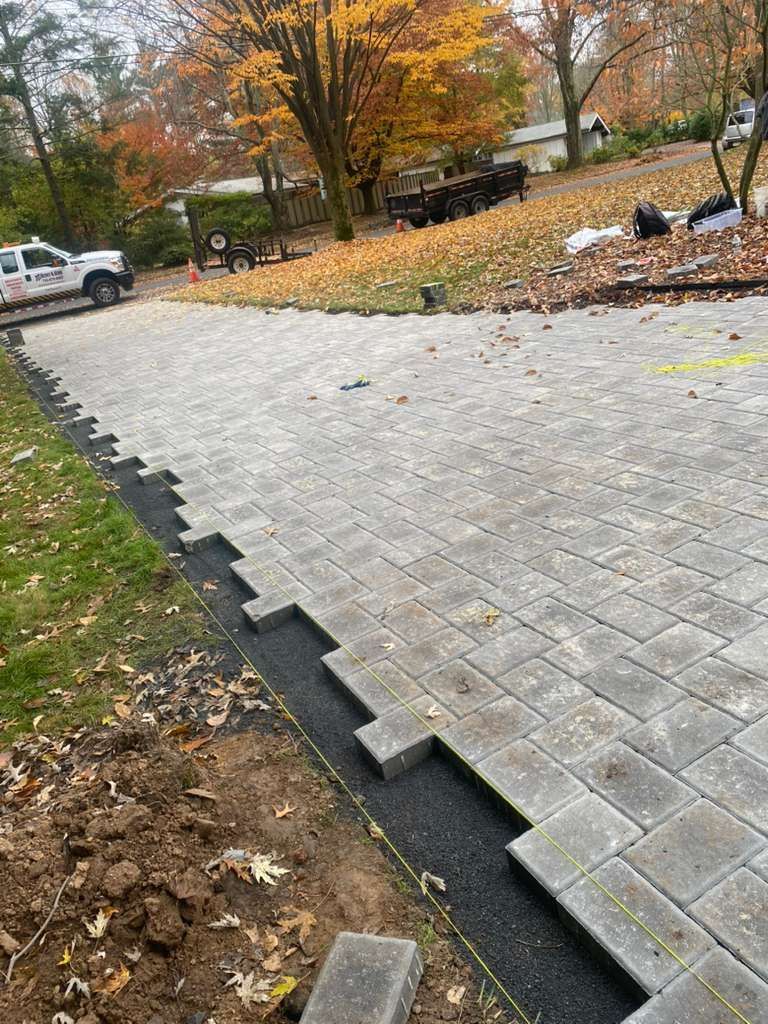 A newly installed light gray paver driveway with a stepped, unfinished edge showing dark gravel base against a lawn.
