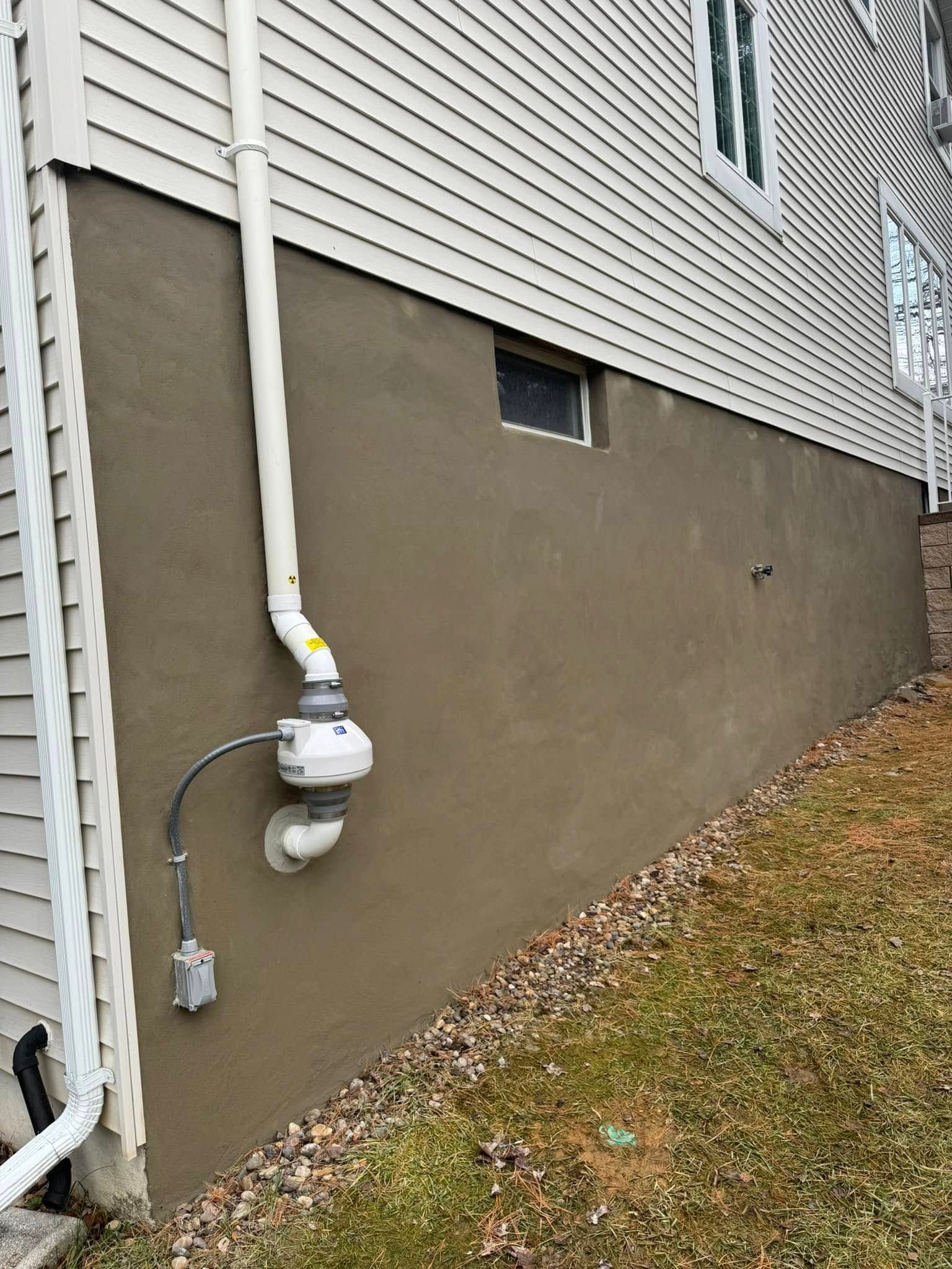 A side view of a house foundation with a radon mitigation system pipe and fan installed on a beige stucco wall.