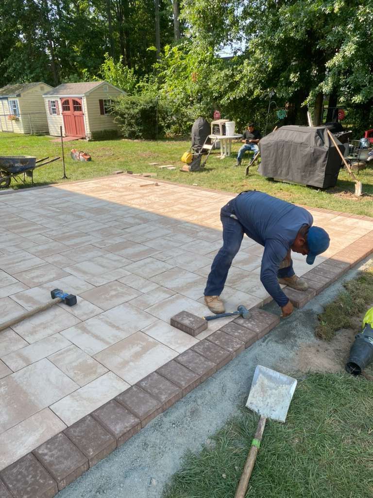 A worker installs light tan pavers with a dark brick border on a patio in a grassy backyard setting.