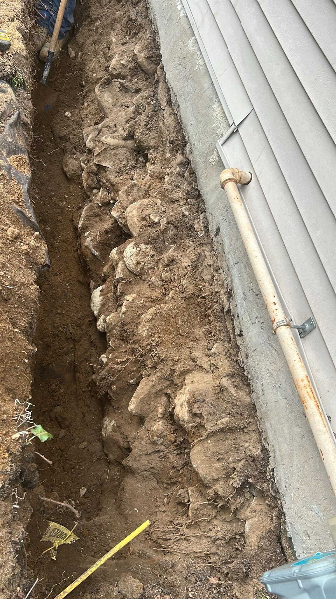 A narrow, deep trench dug in the soil alongside the gray siding and foundation of a house.